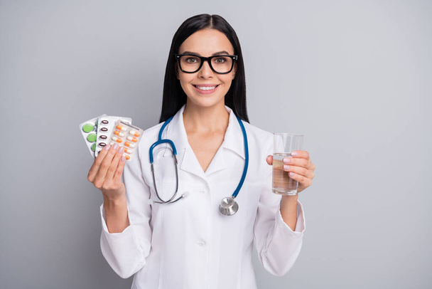 Photo of cheerful doctor girl hold glass water drugs wear specs stethoscope white uniform isolated grey color background - Фото, зображення