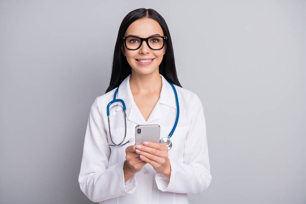 Photo of doctor girl hold smartphone look camera wear stethoscope eyewear white uniform isolated grey color background - Foto, immagini