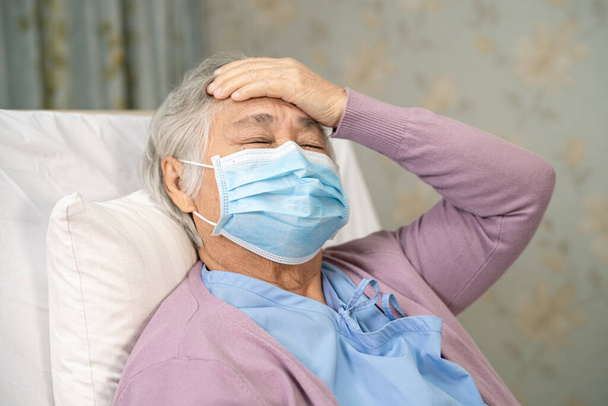 Asyalı yaşlı ya da yaşlı kadın hasta, yüz maskesi takıyor ve baş ağrısı çekiyor. Güvenlik enfeksiyonunu korumak için hastanede yeni bir normal.. - Fotoğraf, Görsel