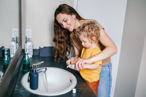 Νεαρή λευκή μητέρα που βοηθάει το παιδί να βουρτσίσει τα δόντια του στο μπάνιο στο σπίτι. Υγιεινή και πρωινή ρουτίνα για τα παιδιά. Η μαμά βοηθάει να μάθει το παιδί να χρησιμοποιεί οδοντόβουρτσα. - Φωτογραφία, εικόνα
