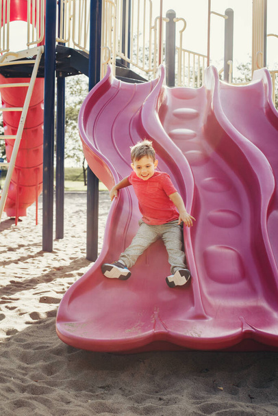 Ativo feliz sorrindo menino caucasiano deslizando no playground. Criança no pátio da escola ao ar livre no dia ensolarado de verão. Criança se divertindo lá fora no campo de jogos. Esportes sazonais atividade infantil. - Foto, Imagem