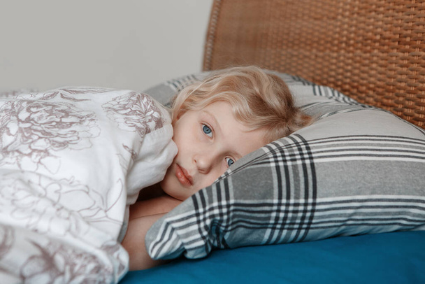 Pensive sad little Caucasian blonde child girl lying on pillow in bed at home. Child lying with open eyes thinking or dreaming. Candid authentic home life. Sweet dreams and bed vibes.  - Photo, Image