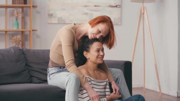 roodharige vrouw knuffelen hispanic vriendin en het vasthouden van handen in de woonkamer - Video