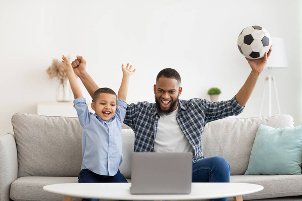 African Father And Son Watching Soccer Online At Laptop Indoors - 写真・画像