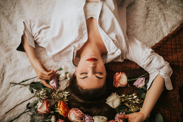 Μια γυναίκα με λευκό πουκάμισο ξαπλώνει σε ένα κρεβάτι με λουλούδια. Δώρο του Αγίου Βαλεντίνου. Γυναικείο δώρο, 8 Μαρτίου. - Φωτογραφία, εικόνα