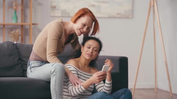 διαφυλετικό λεσβιακό ζευγάρι κοιτάζοντας smartphone στο σαλόνι  - Πλάνα, βίντεο