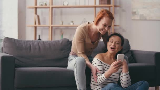 πολυπολιτισμικό λεσβιακό ζευγάρι κοιτάζοντας smartphone και χαμογελώντας στο σαλόνι  - Πλάνα, βίντεο