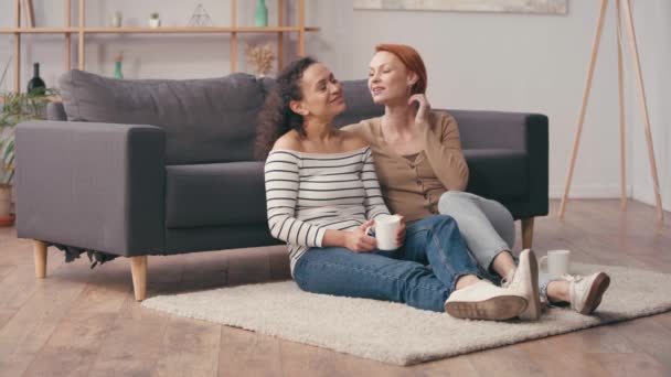 roodharige vrouw knuffelen hispanic vriendin met kopje in woonkamer - Video