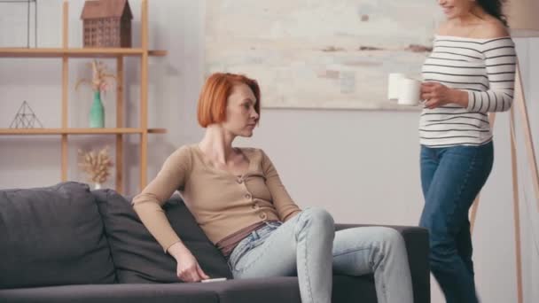 Spanierin schenkt glücklicher Freundin mit Smartphone Tasse im Wohnzimmer - Filmmaterial, Video