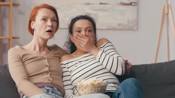 zszokowana wielokulturowa lesbijka ogląda straszny film w pobliżu miski popcornu  - Materiał filmowy, wideo