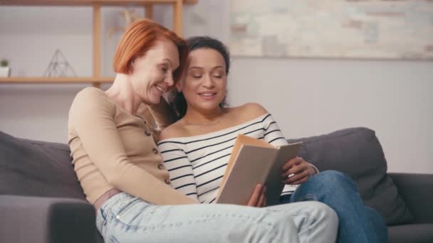 hispanic woman holding book and talking with cheerful girlfriend in living room - Footage, Video