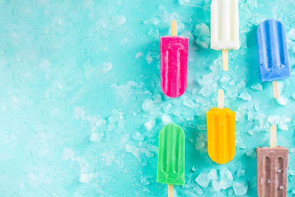 Selection of bright multicolored ice cream popsicle. Various gelato, frozen lollypops - chocolate vanilla blueberry strawberry pistachio orange, with crushed ice on light blue sunny background - Photo, Image