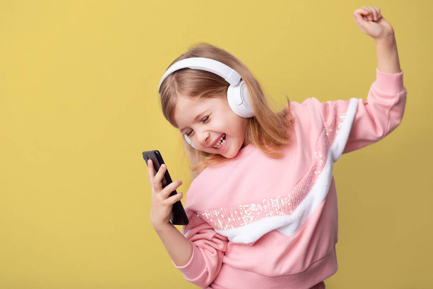 A little girl blogger influencer records a blog video on a smartphone, dances and listens to music with headphones on a yellow isolated background. - Фото, изображение