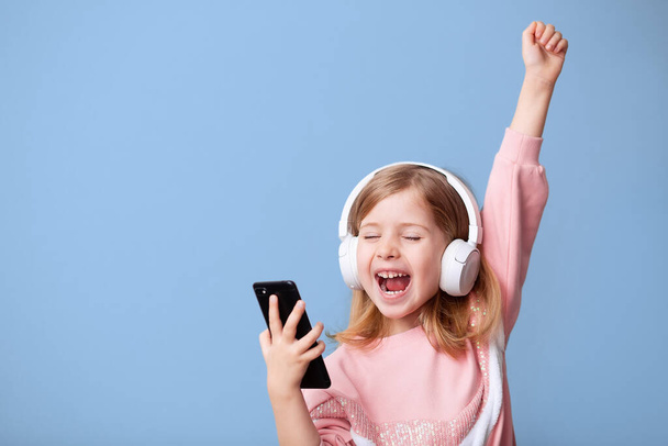 A little girl-blogger influencer records a video from a blog on a smartphone, dances, listens to music with headphones, rejoices in victory on a blue isolated background. - Photo, Image