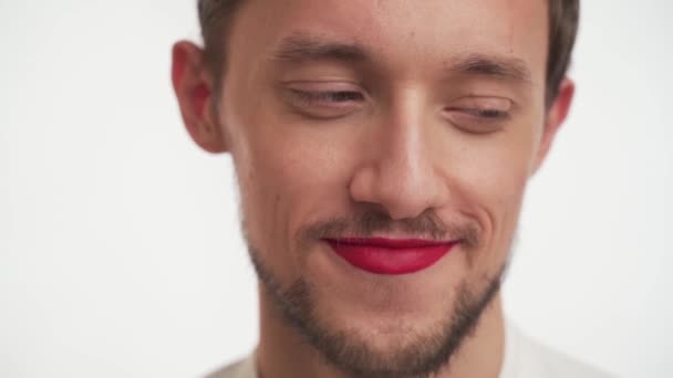 Young cute transvestite embarrassed brunette man with dark black beard, moustache, painted lips in red lipstick rolls eyes, looks around, stare at camera, smiles isolated on white background close up. - Footage, Video