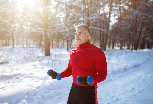 Outdoor wintersport concept. Sterke senior man die traint met halters, zijn spieren traint in het snowy park - Foto, afbeelding