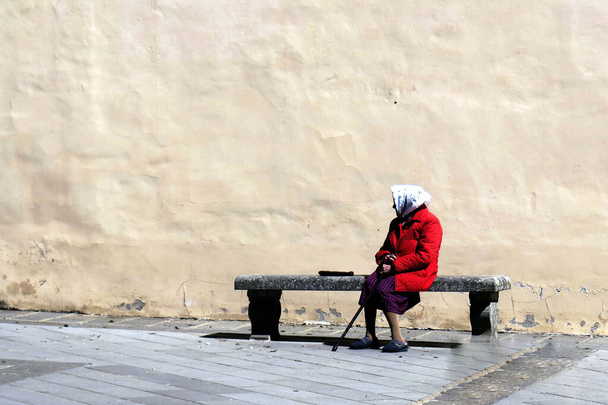 een oudere dame zit alleen op een bank met de muur van een oud gebouw op de achtergrond - Foto, afbeelding