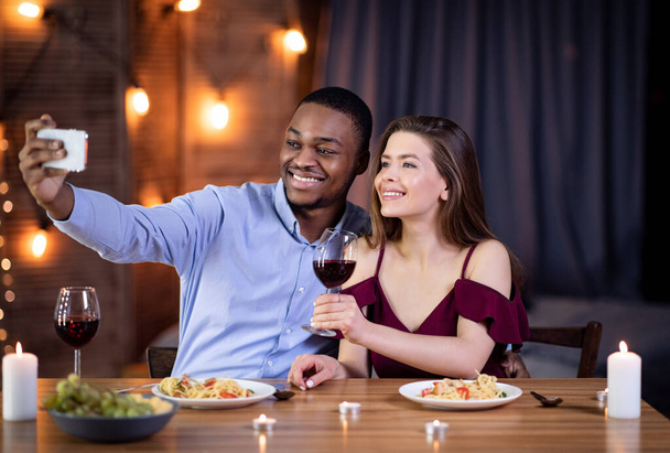 Pareja Interracial feliz tomando selfie en Smartphone durante la cita romántica en el restaurante - Foto, Imagen