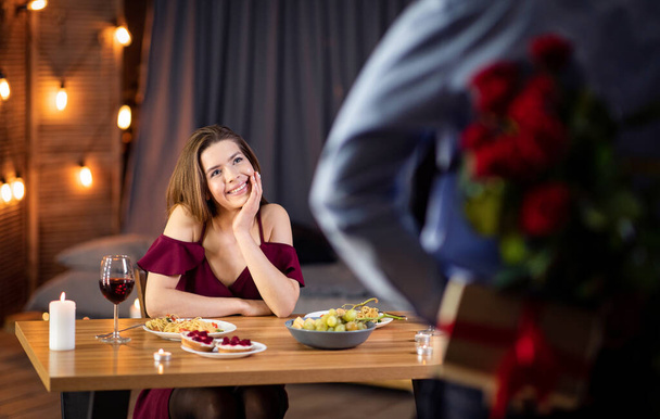 Hombre haciendo sorpresa romántica a su novia en el restaurante, sosteniendo rosas y regalo - Foto, imagen