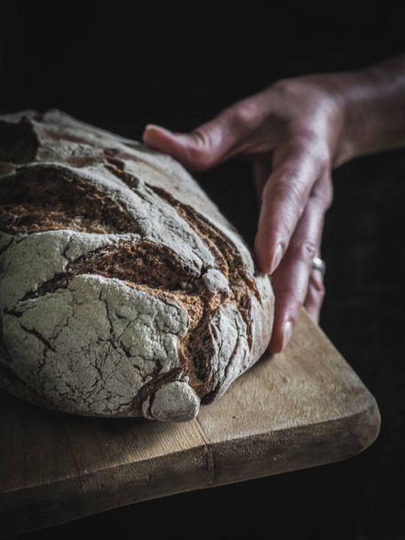 Θηλυκό χέρι κρατά καρβέλι ψωμί σε ξύλινη επιφάνεια κοπής. Σκοτεινός και κυκλοθυμικός. Αντιγραφή space.f - Φωτογραφία, εικόνα