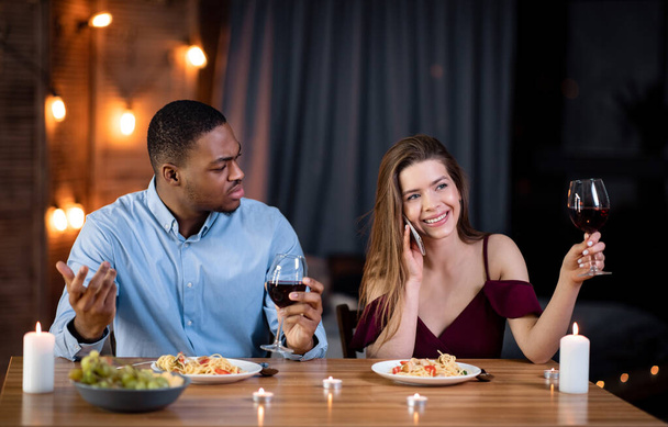 Femme parlant sur téléphone portable pendant un rendez-vous romantique, assis à côté de son petit ami en colère - Photo, image