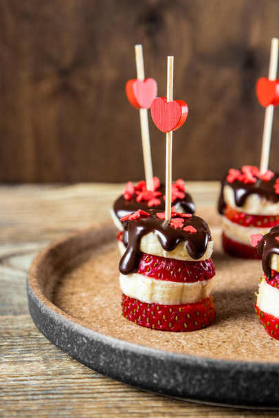  Banana and strawberry canapes in chocolate glaze on a plate, an original appetizer for Valentine's Day, vertical photo. High quality photo - Φωτογραφία, εικόνα