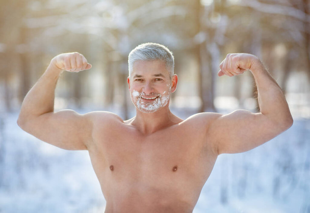 sportif senior avec de la neige sur le visage et le torse nu montrant des muscles biceps proéminents à l'extérieur par temps froid d'hiver - Photo, image