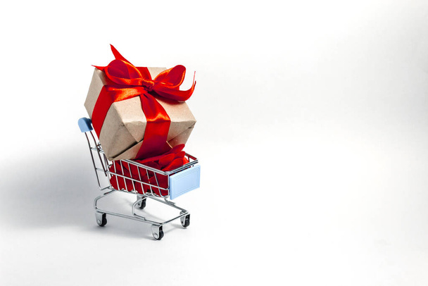 A supermarket cart filled to the brim with red confetti hearts and a gift box wrapped in kraft paper and tied with a red ribbon on a white background. The concept of buying presents. Copy space. - Photo, image
