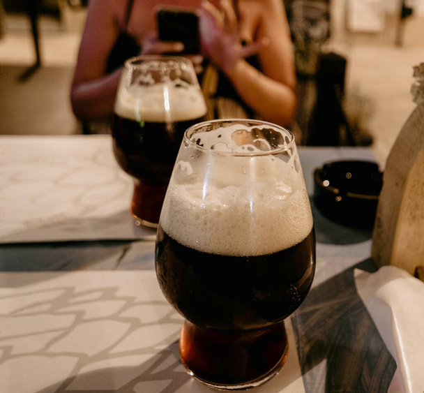 Selective focus close-up image of beer glasses on table in bar. Date night, woman in background, craft beer, drinks, alcohol. - 写真・画像