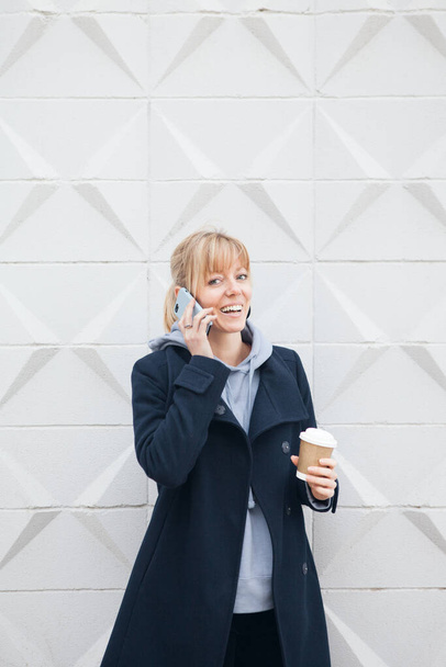 Neşeli sarışın, yüzü memnun olmuş Avrupalı bir kadın, cep telefonuyla konuşuyor, gezinti bağlantısını kullanıyor, nefis bir kahve tutuyor. İletişim konsepti - Fotoğraf, Görsel