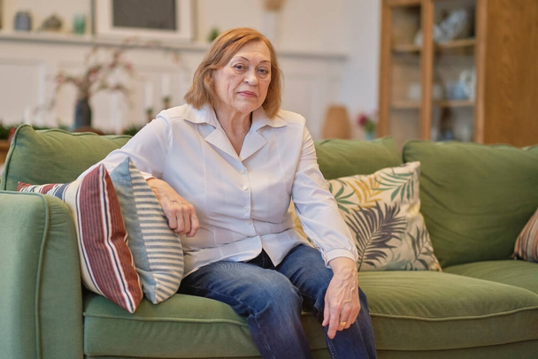 Ritratto di una donna anziana seduta su un divano in una casa di riposo. Donna caucasica seduta su un divano e che guarda la telecamera. Cura dei pensionati soli. - Foto, immagini