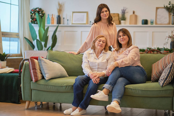 three generations of a family of women are sitting together on the couch. grandmother, daughter, and granddaughter pose together to get a joint photo. - Photo, Image
