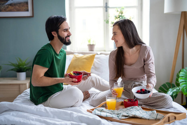 Young couple in love eating breakfast on bed indoors at home, talking. - Фото, зображення