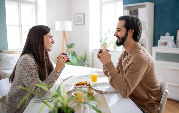 Young couple in love eating hamburgers indoors at home. - Photo, Image