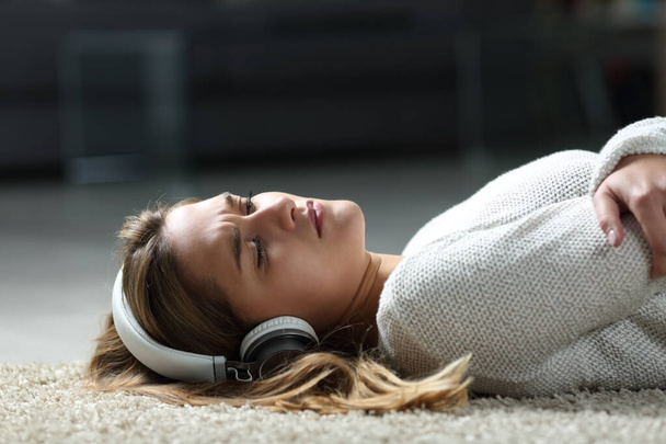 Sad woman listening to music wearing headphones lying on the floor at home - Foto, afbeelding