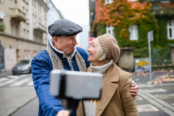 Happy senior couple walking outdoors on street in city, taking selfie. - Фото, изображение