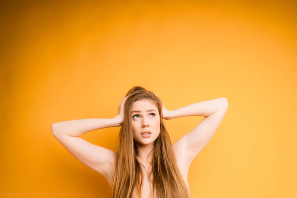 Attractive Caucasian girl holds her head with her hands and is surprised on an orange background. Space for text - Φωτογραφία, εικόνα