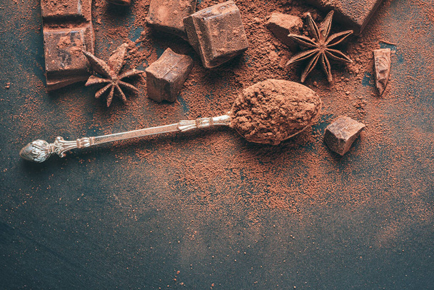 Brown cocoa powder in the spoon and chopped chocolate cubes top view on dark background with copy space. - Fotó, kép