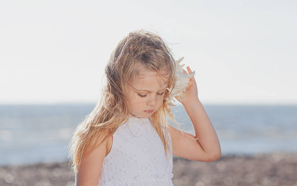 Μικρό καυκάσιο ξανθό κορίτσι με λευκό φόρεμα που κρατάει κοχύλι και ακούει τη θάλασσα - Φωτογραφία, εικόνα