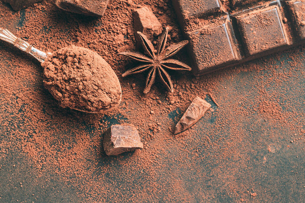 Brązowy proszek kakaowy w łyżce, batonik czekoladowy i posiekane kostki czekolady, widok z góry na ciemne tło z przestrzenią do kopiowania. - Zdjęcie, obraz