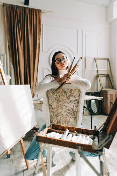 Portrait of beautiful smiling woman artist, sitting on a chair and holding paintbrushes, working on a home studio. - Fotografie, Obrázek