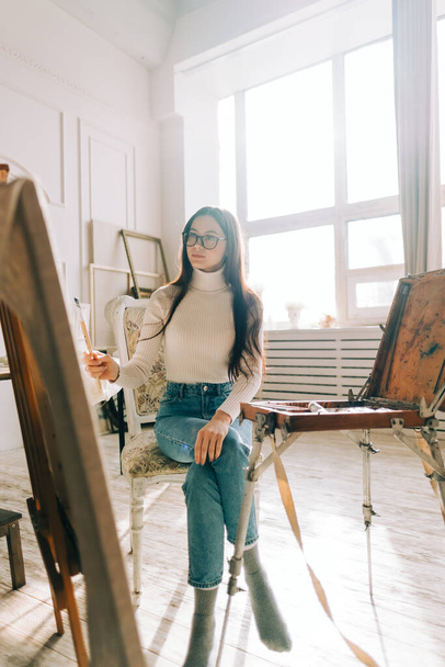 Young caucasian woman artist, holding paintbrush and draws on canvas , working on a home studio. - 写真・画像