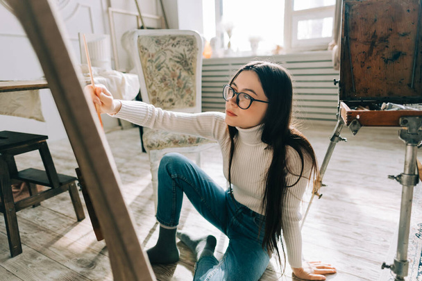 Young caucasian woman artist, sitting on a floor, holding paintbrush and draws on canvas , working on a home studio. - Foto, immagini