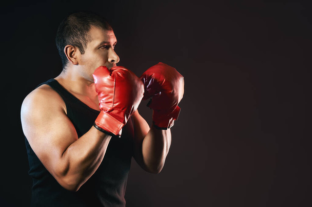 El luchador masculino hispano está boxeando y entrenando con guantes de boxeo y está usando una camiseta que muestra sus brazos fuertes y musculosos. Aislado sobre fondo negro con espacio de copia - Foto, Imagen