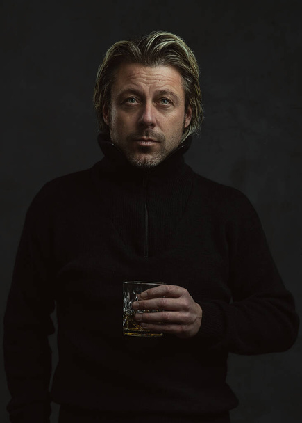 Homme blond en pull en laine noire tenant un verre de whisky. - Photo, image