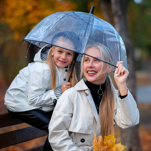 Mom and daughter are sitting in the park on a bench under a transparent umbrella. Autumn park, family day - Фото, изображение