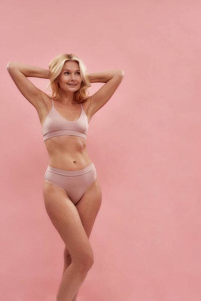 Feeling beautiful at any age. Happy attractive middle aged caucasian woman in underwear smiling and looking away while standing against pink background, vertical shot - 写真・画像