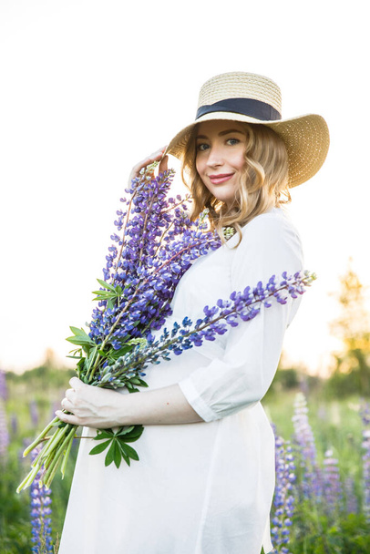 a young beautiful girl in a white dress and hat walks across the field with lupins - Foto, afbeelding