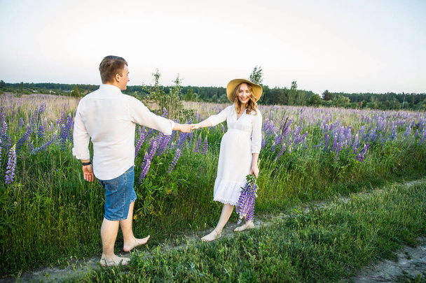 young beautiful couple a girl is pregnant in a white dress and a hat a man in a white shirt are walking along the field with lupins at sunset - Fotografie, Obrázek
