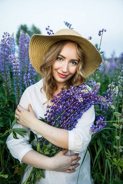 a young beautiful girl in a white dress and hat walks across the field with lupins - Фото, изображение